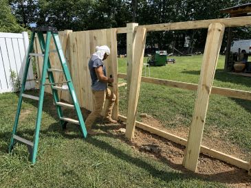 Person building a wooden fence in a backyard using a ladder.