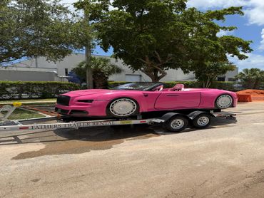 Bright pink convertible car on a trailer under a sunny sky.