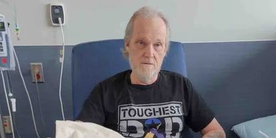 Elderly man wearing a 'Toughest Dad' shirt sitting in a hospital bed.