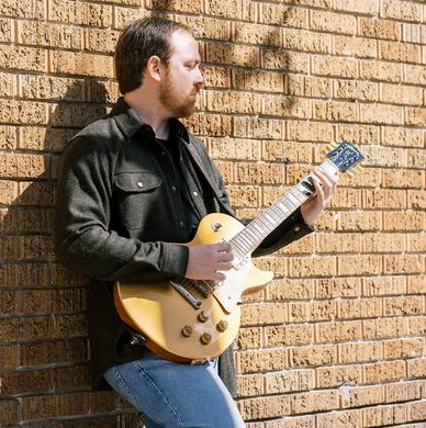 Man playing electric guitar against a brick wall.