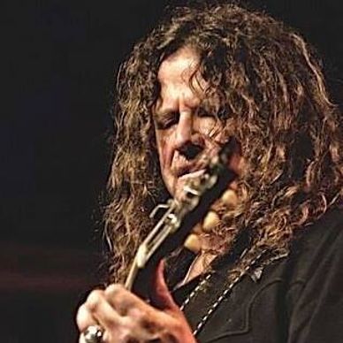 Man intensely playing guitar on stage with long curly hair.
