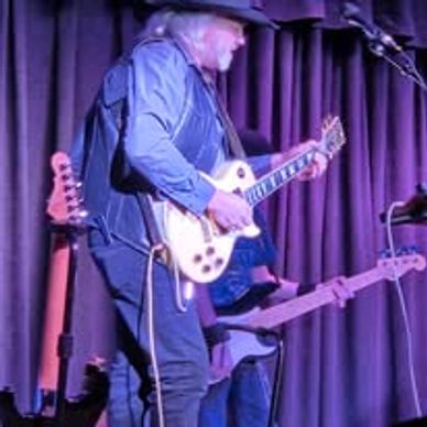Musician playing electric guitar on stage with a purple curtain backdrop.