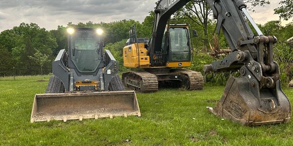 Two construction machines, a skid steer loader and an excavator, on green grass.