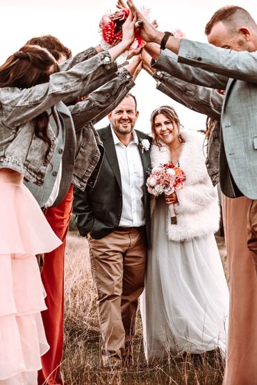 Newlyweds smile under a joyful arch of raised hands and flowers at their wedding.