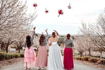 Bride and bridesmaids toss bouquets in a blossom-lined outdoor setting.
