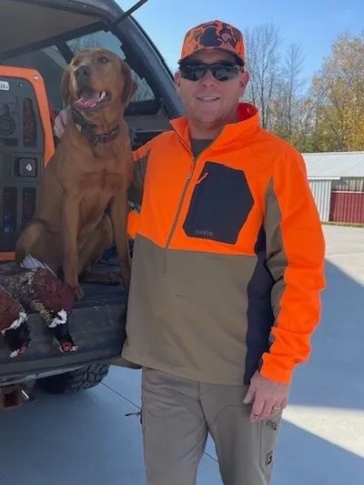 Man in hunting attire with a dog and pheasants in a vehicle.