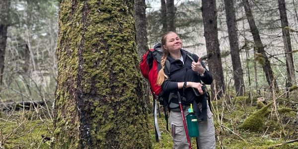 Woman hiker giving thumbs up in a mossy forest with backpack and hiking poles.