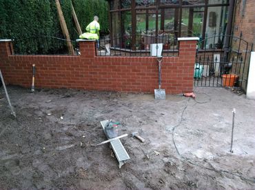 Garden landscaping work in progress with tools and worker near a brick wall.