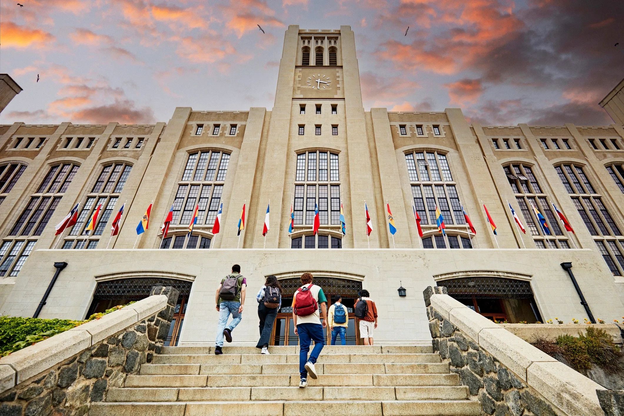 Students walk up steps toward a grand university building at sunset.