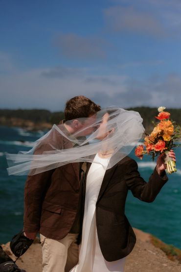 Couple sharing a kiss by the ocean with a flowing veil and bouquet.