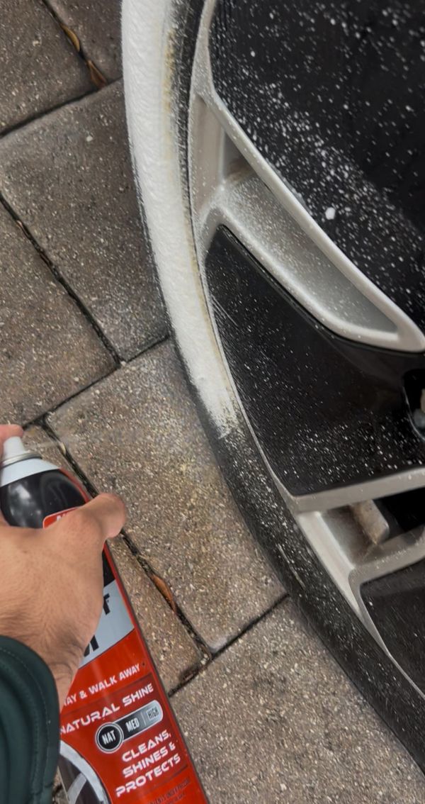 Hand spraying foam cleaner on a car wheel and tire on a paved surface.