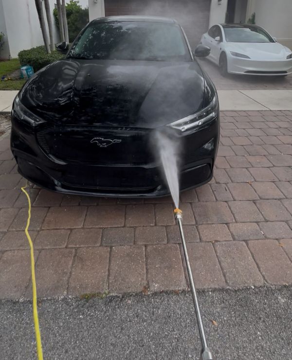 A black Mustang being pressure washed in a driveway with a white Tesla in the background.