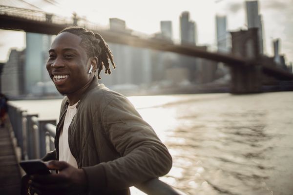Man standing on the river edge in a city talking on the phone (with ear buds) and smiling