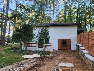 Modern white brick house with wooden accents surrounded by tall trees and a wooden fence.