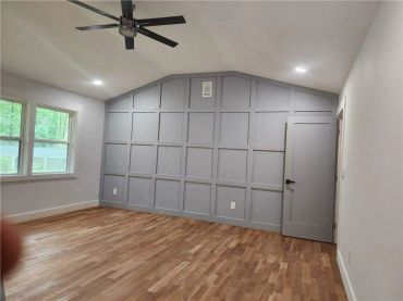 Empty room with wooden floor, gray paneled accent wall, ceiling fan, and windows.