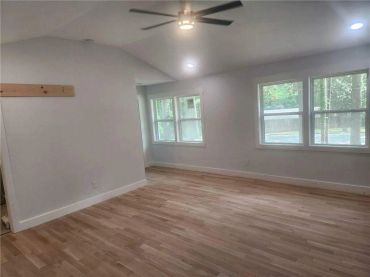 Empty room with wooden floor, white walls, ceiling fan, and windows.