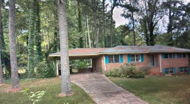 Brick house with teal shutters surrounded by tall trees and greenery.