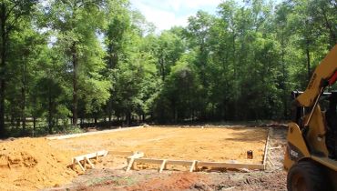 Cleared construction site with wood framing and a backhoe near a forest.