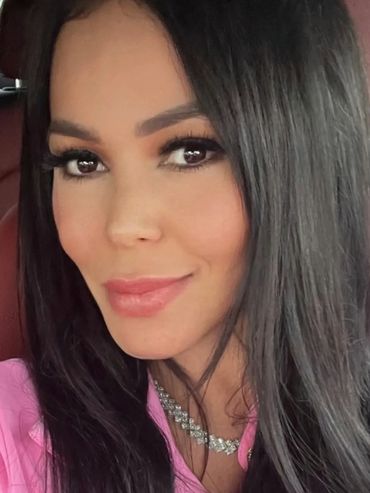 Close-up of a woman with long dark hair, wearing a pink top and a diamond necklace.