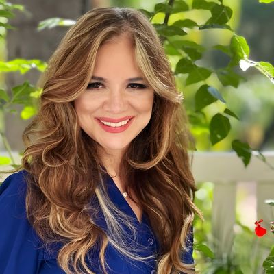 A smiling woman with long wavy hair in a blue blouse stands outdoors near green plants.