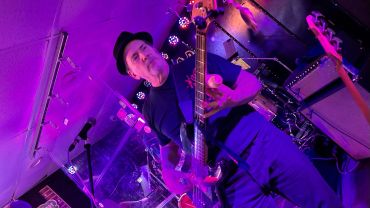 Man playing bass guitar on stage with purple lighting.