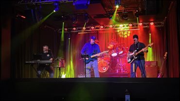 A four-member band performs on stage with colorful lighting and instruments.