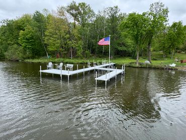 A white dock with chairs and an American flag on a calm lake surrounded by trees.