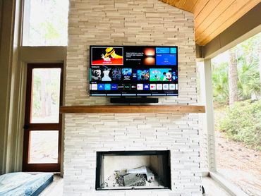 Modern living room with TV and soundbar mounted above a white stone fireplace and large windows. Hilton Head Island, SeaPines neighborhood, brand newly constructed home, installed and supported by The Gadget Tutor Tech Support and Installers.