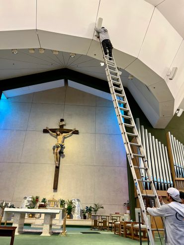 A worker on a tall ladder inside a church near a crucifix and altar.