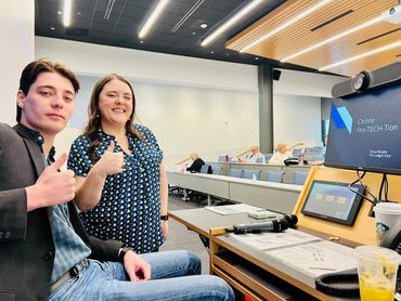 Two people giving thumbs up in a modern classroom with a presentation screen.