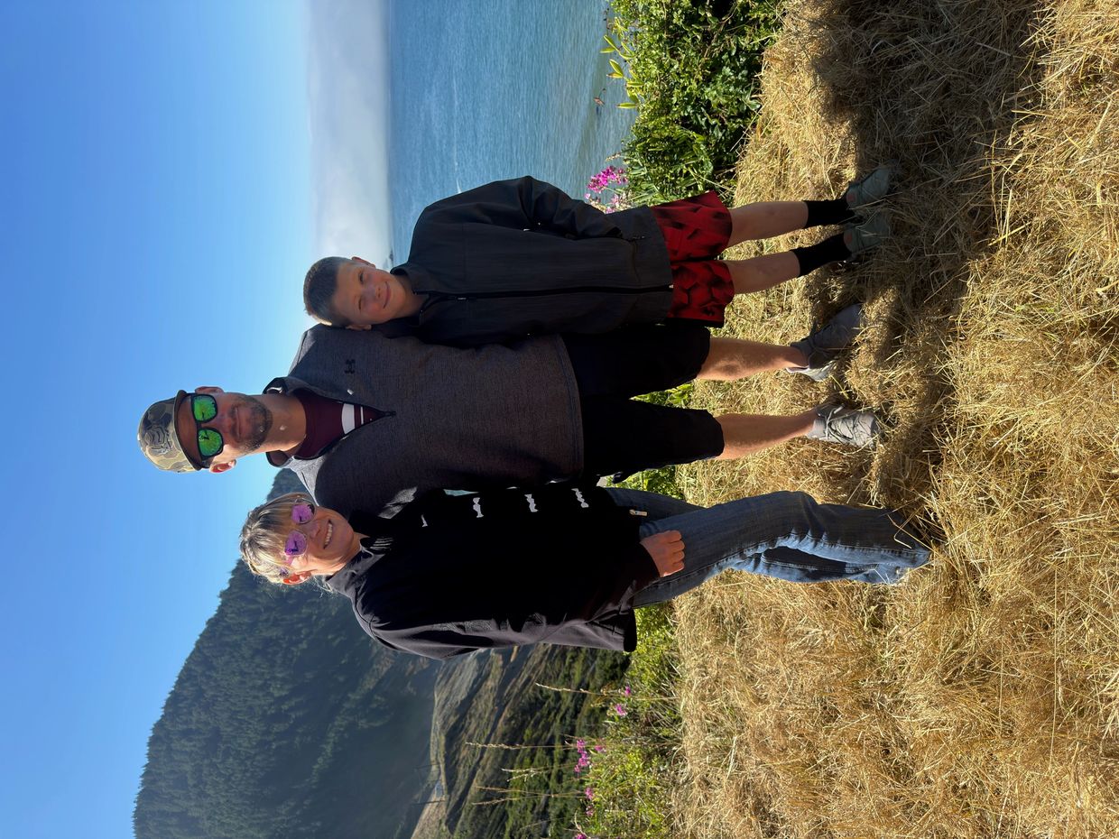 A family of three posing outdoors near a body of water on a sunny day.