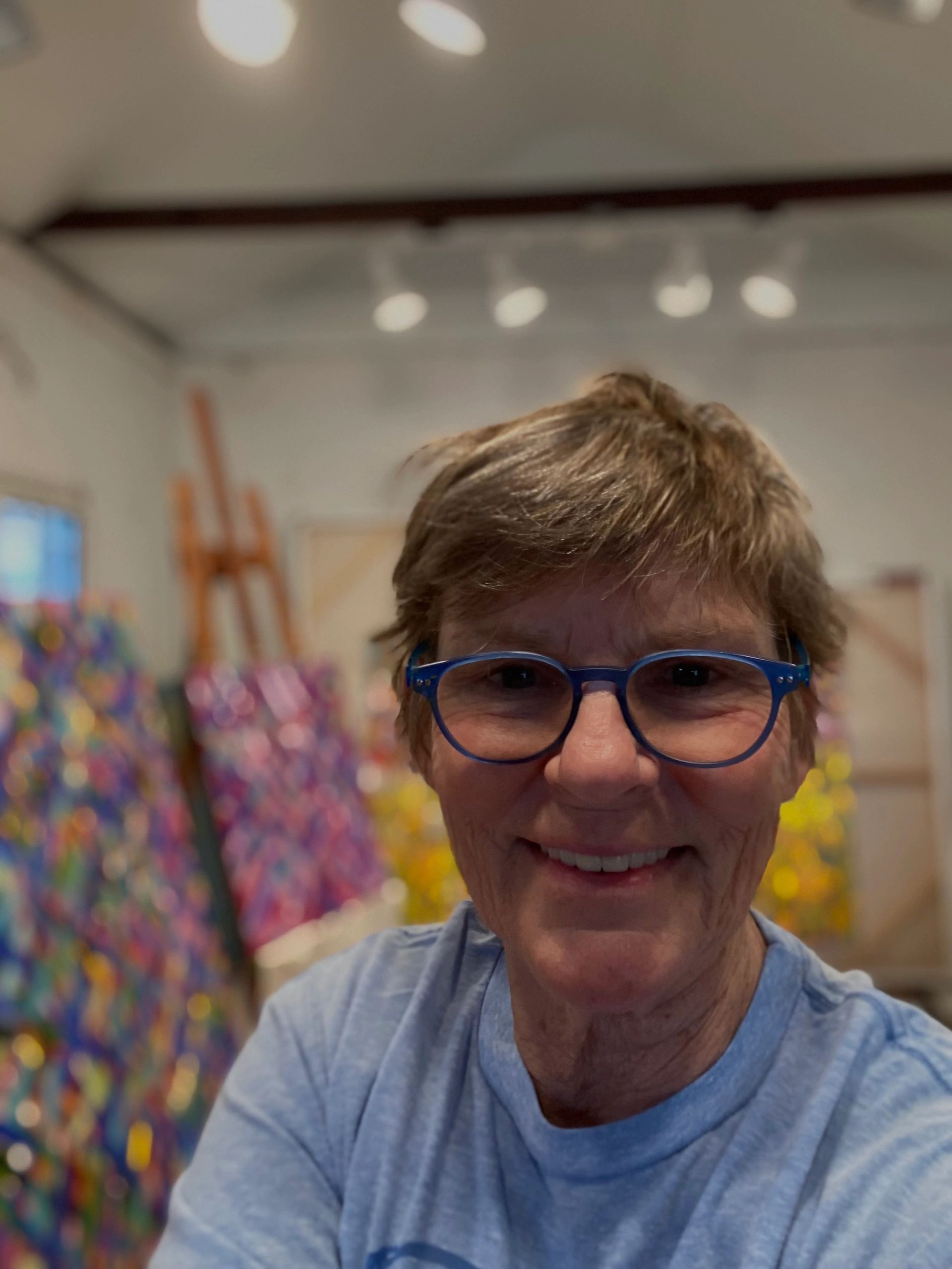 Smiling woman with blue glasses in an art studio with colorful paintings.