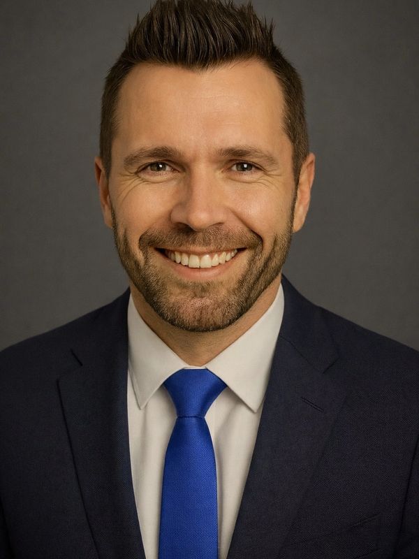 Smiling man in a navy suit and blue tie against a gray background.
