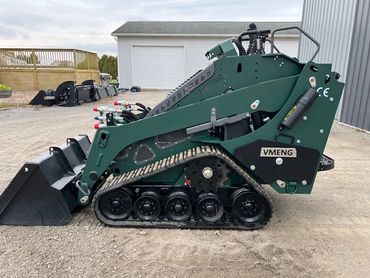 Compact green VMENG tracked loader parked on gravel near a building.