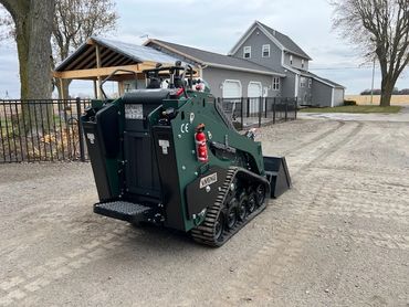 Compact tracked loader parked on a gravel driveway near a residential house.