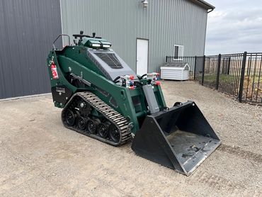 Compact green skid steer loader with bucket attachment on gravel near building.