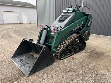 Compact green tracked loader with a front bucket attachment on a gravel surface.