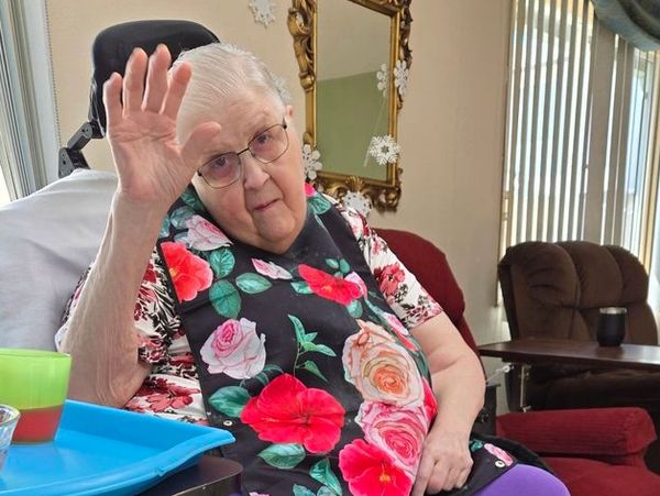 Elderly woman waving while sitting indoors.