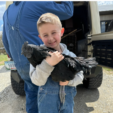 A boy happily holding a black chicken outdoors near a vehicle.