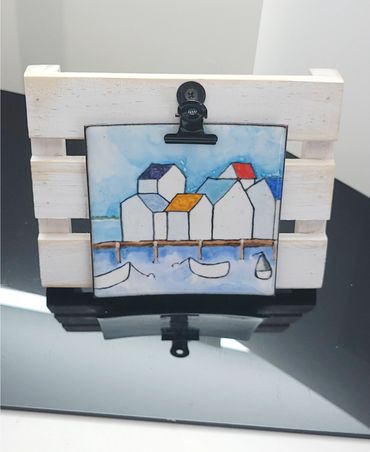 Small hand painted enamel tile with painted houses on a dock, held by a clip on a wooden stand. Siz