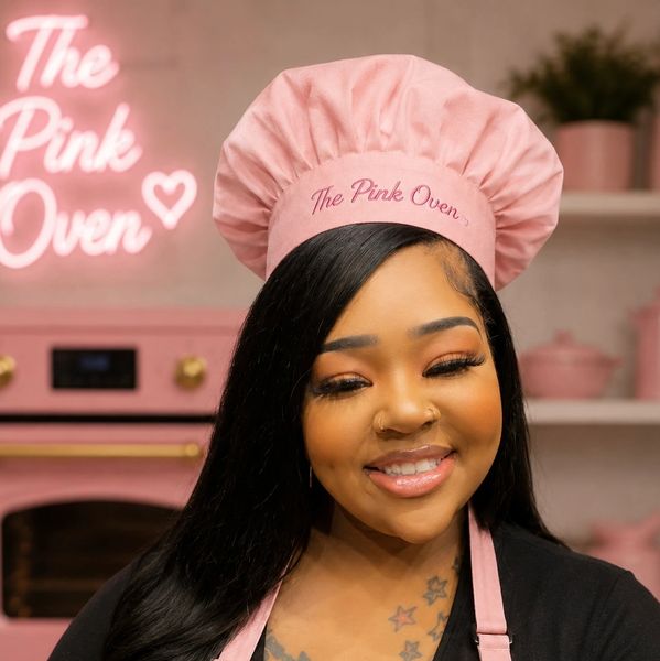 Smiling woman wearing a pink chef hat and apron in a pink kitchen.