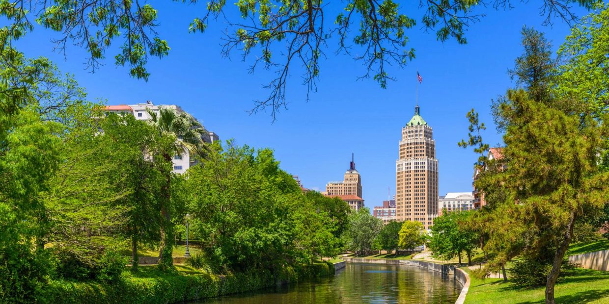 A river flows through a green park with city buildings in the background under a clear blue sky.