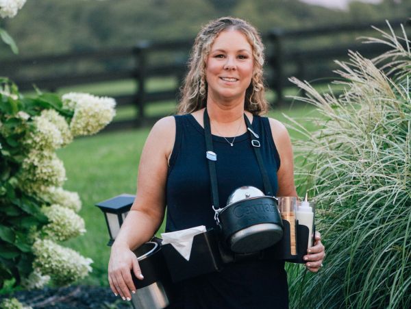 Woman smiling outdoors with a drink carrier strapped around her neck.