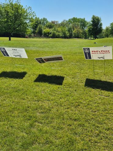 Three small advertising signs on a grassy field with trees in the background.