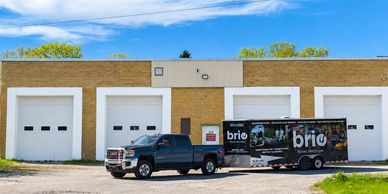 A black pickup truck with a branded trailer parked in front of a warehouse with white garage doors.