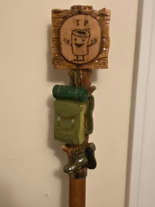 A decorative walking stick with camping-themed carvings and a smiling toilet paper sign.