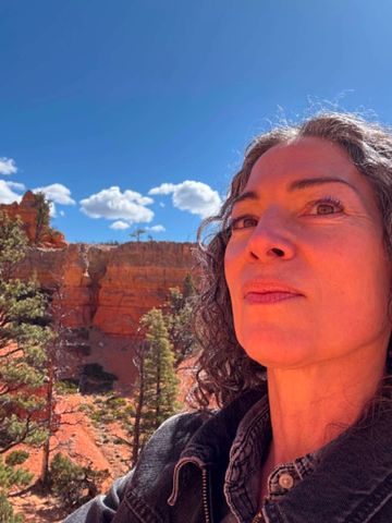 Woman with curly hair stands in front of a red rock canyon under a blue sky.