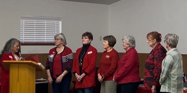 A woman speaks at a podium to six women standing in a row indoors.