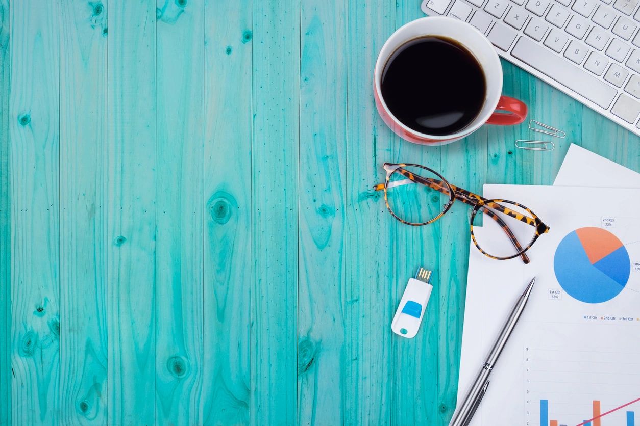 A green desk with a cup of coffee, computer keyboard, phone charger, glasses and business papers.