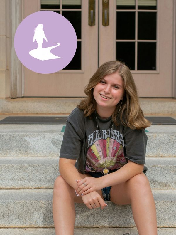 Young woman sitting on stone steps, smiling, wearing a dark graphic t-shirt. Her Path Forward